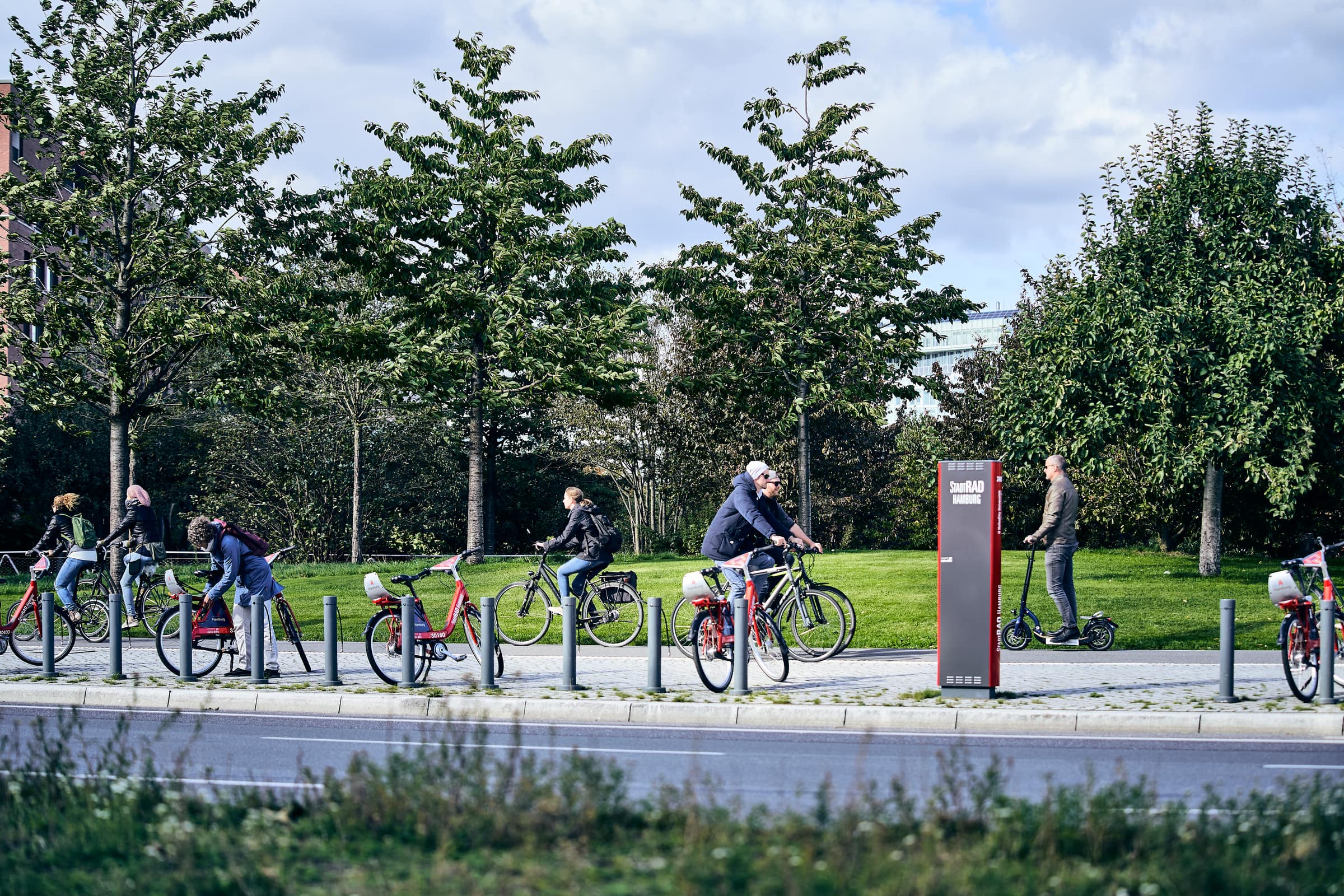 Personen fahren auf Fahrrädern und Rollern, dahinter liegt der Lohsepark, davor ist eine Straße mit Stadtrad-Station